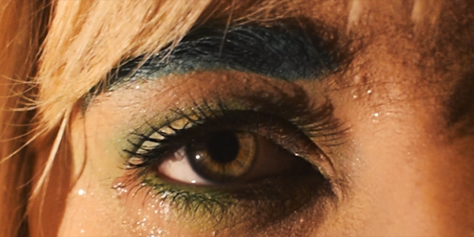 A close up of a hazel-coloured eye, with green eyeshadow and black eyebrows, and blonde hair.